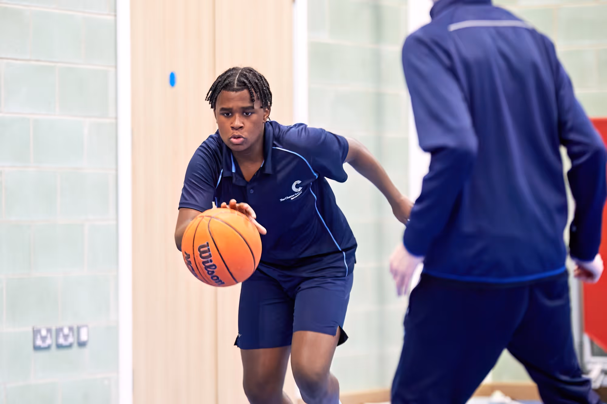 Teen boy in navy sportswear dribbling a basketball indoors while guarded by another player.