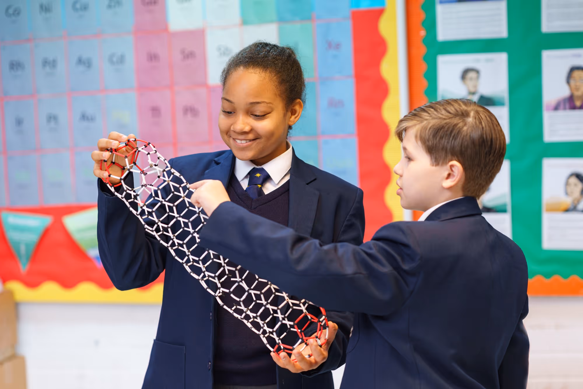 Two students in school uniforms holding and examining a geometric molecular model in a classroom.