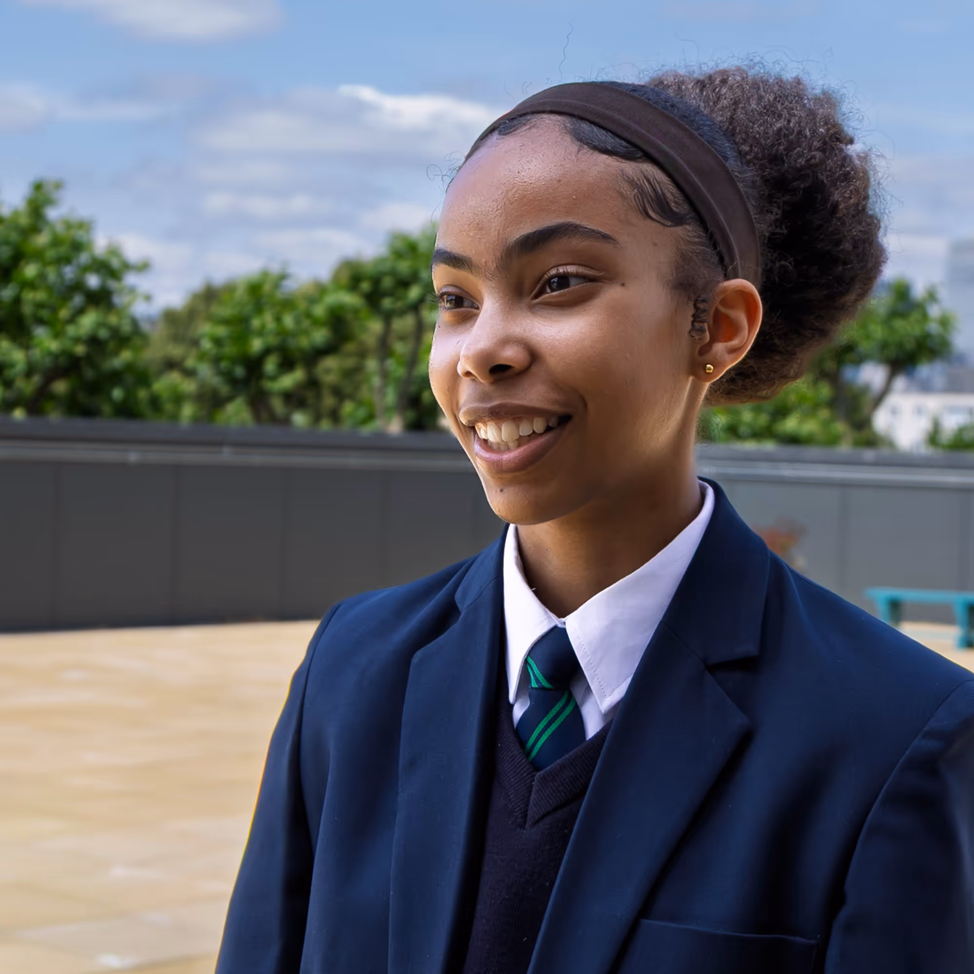 Smiling girl in a school uniform with blazer and tie standing outdoors in daylight.