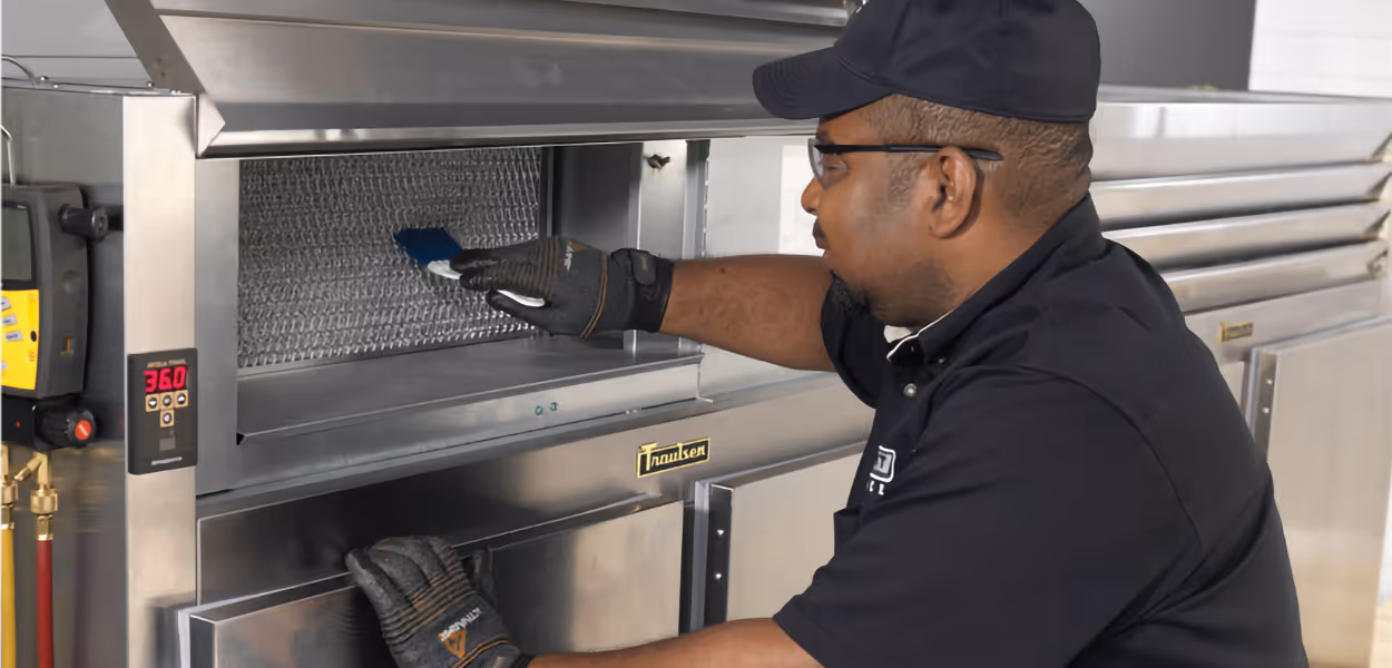 Technician wearing gloves and a cap cleaning the interior of a commercial refrigerator unit.