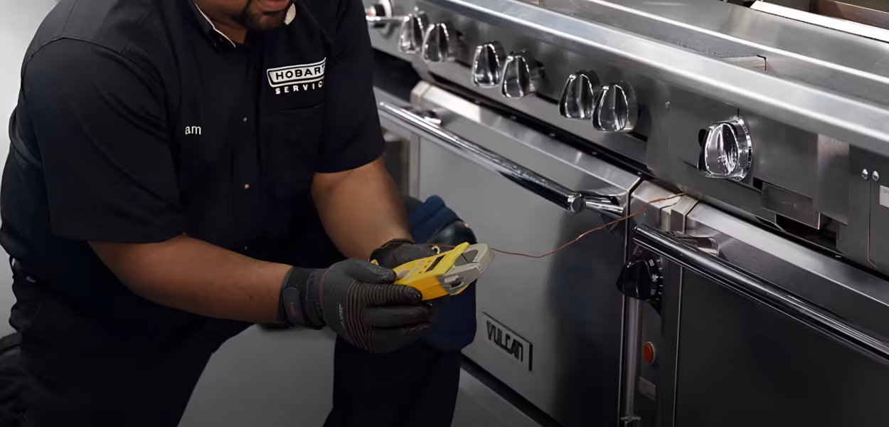 Technician wearing gloves using a yellow diagnostic device to repair a commercial stainless steel Vulcan stove.