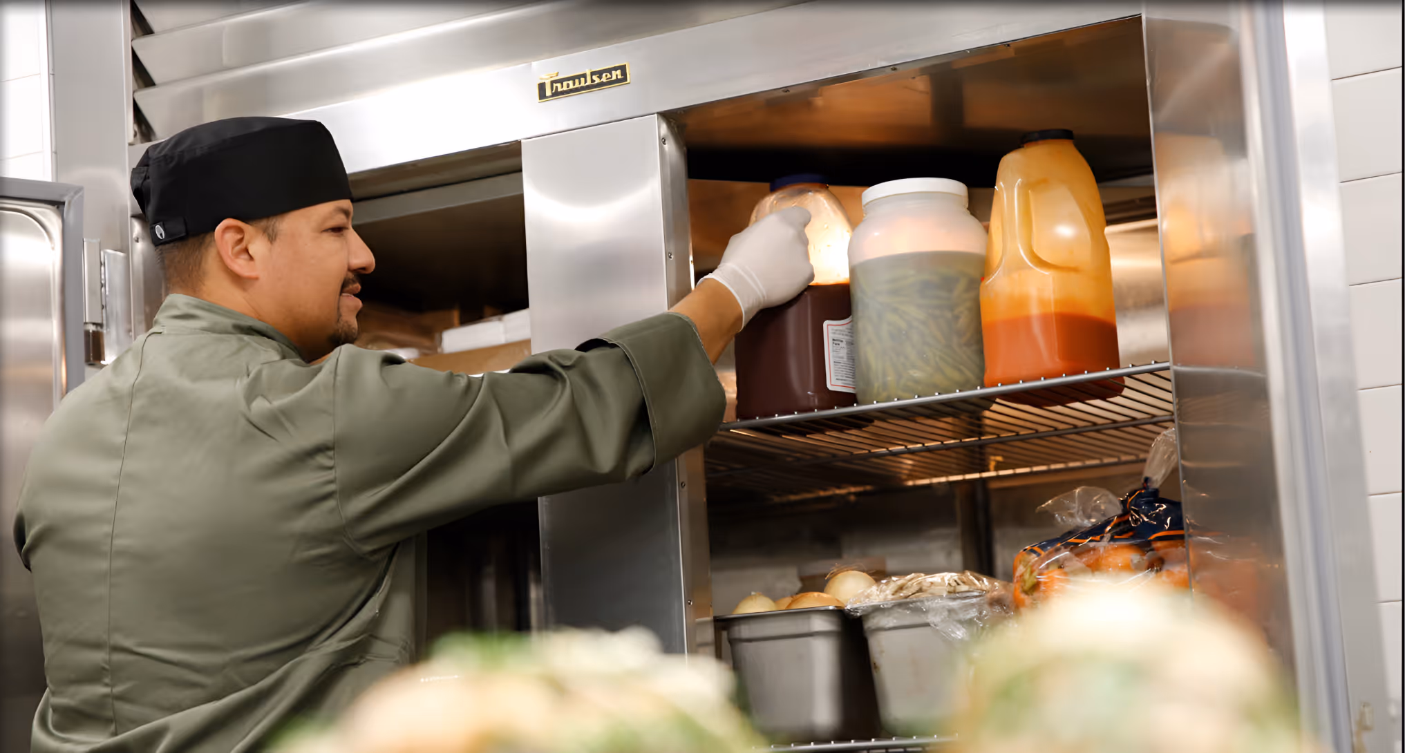 Chef in green uniform and black hat reaching for a container inside a commercial kitchen refrigerator.