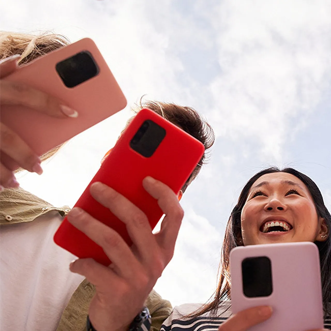 Three people smiling and holding smartphones with colorful cases against a cloudy sky.
