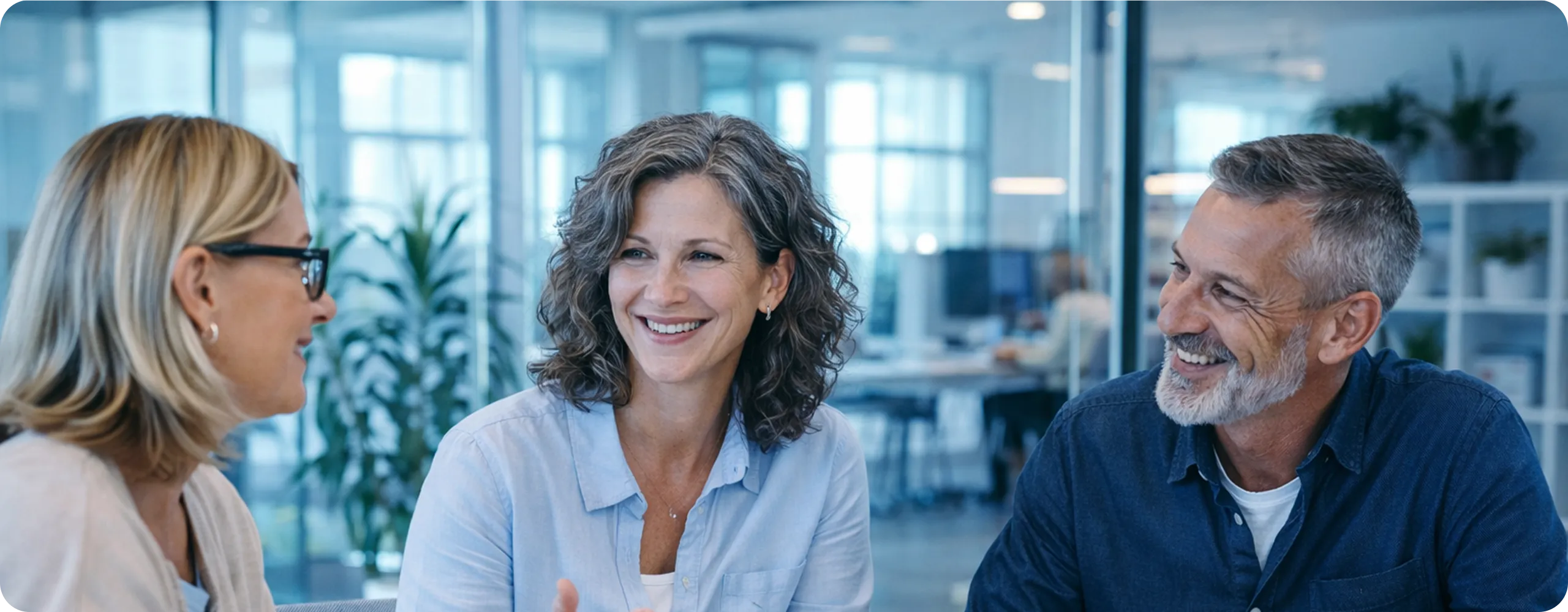 Three colleagues smiling and having a conversation in a modern office setting.