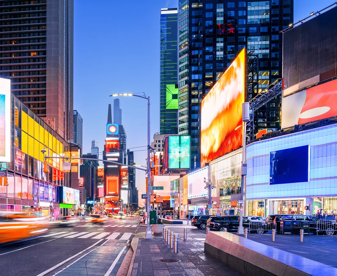 Busy New York City street at dusk, with illuminated billboards, tall buildings, traffic, and Times Square-style city activity.