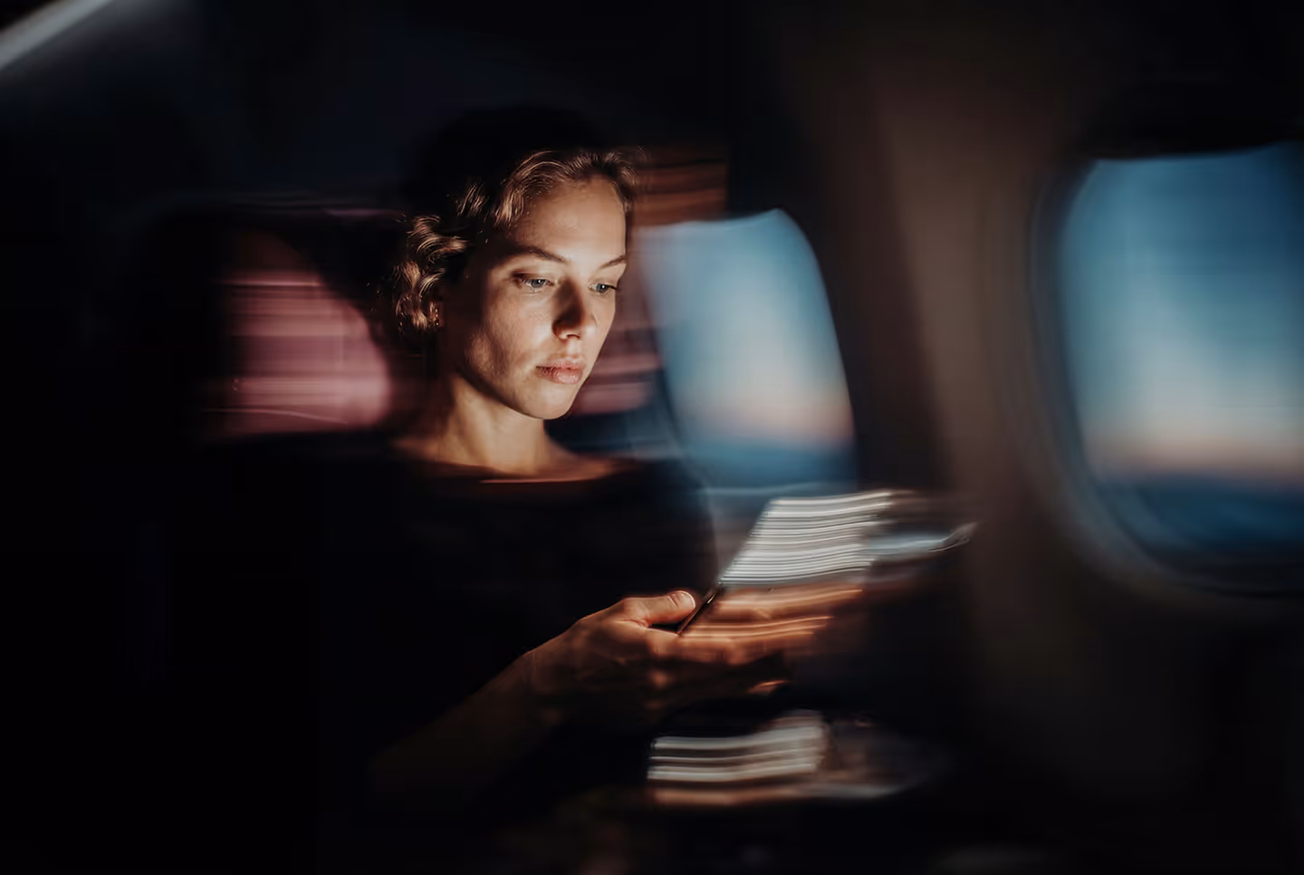 Woman sitting on an airplane looking at her smartphone with light trails around her in dim cabin light.