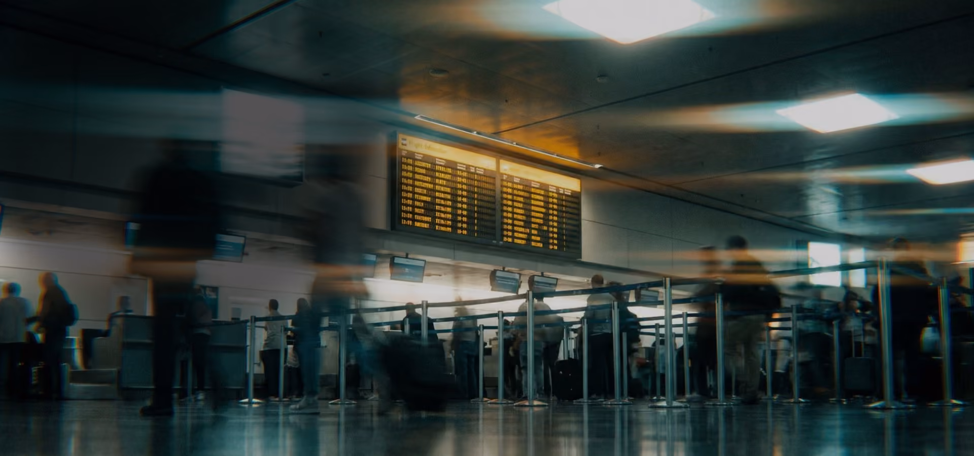 Passengers with luggage waiting in line at an airport check-in counter under a flight info board.