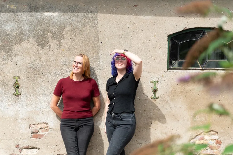 Foto van Jonneke en Odile in het zonnetje voor een oude muur. 