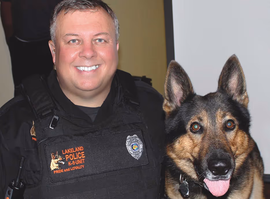 Smiling police officer in black Lakeland Police K-9 Unit uniform with a German Shepherd dog.