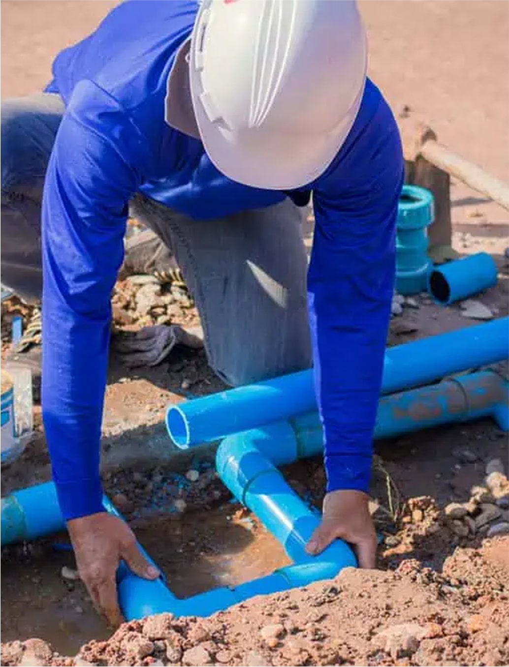 Worker in protective gear installing blue pipe underground.