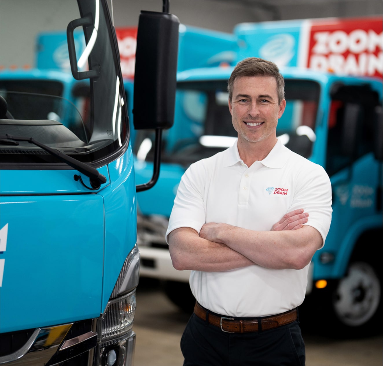 Technician in white shirt with arms crossed standing near service trucks.