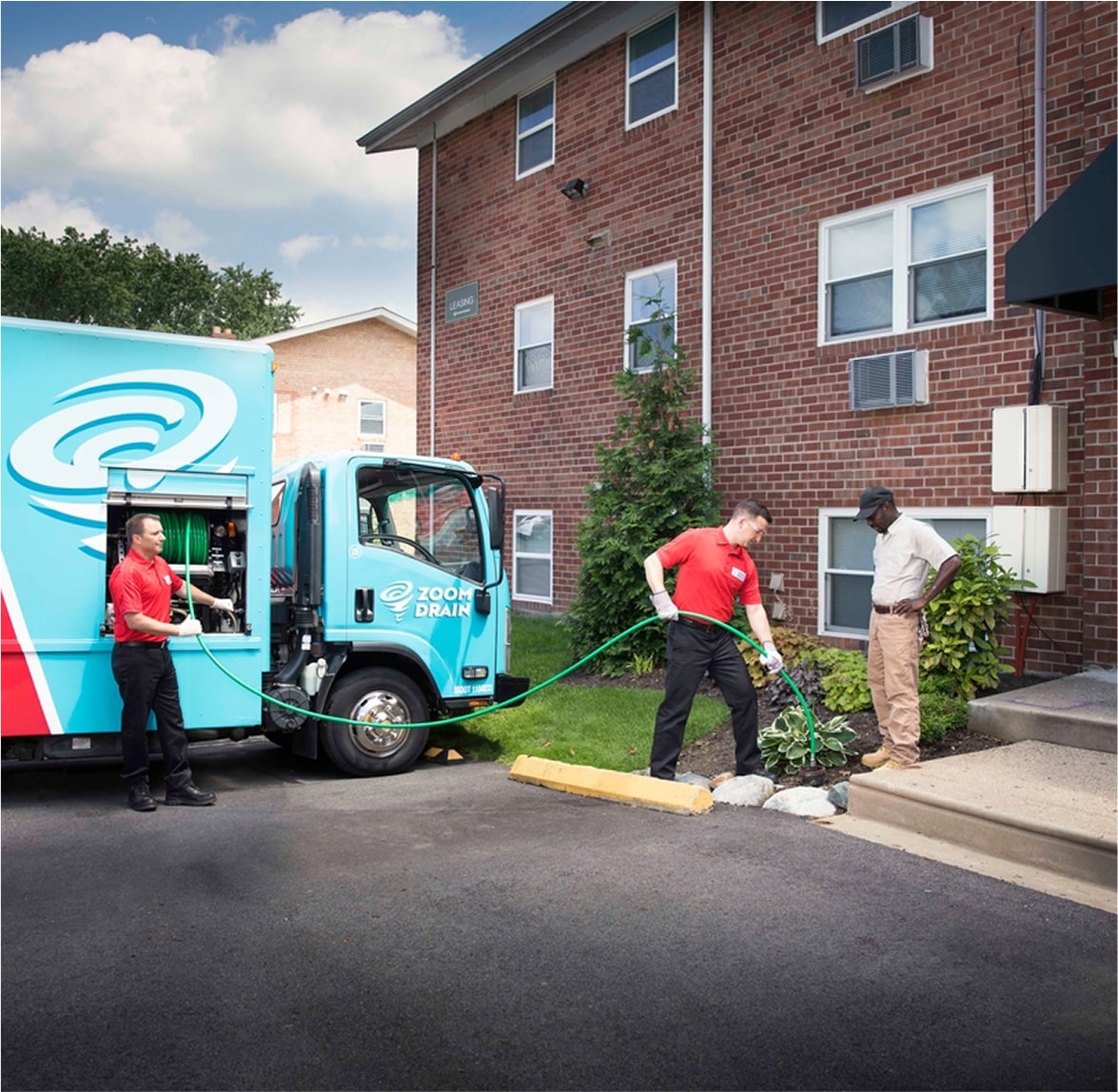 Two technicians working outside a residential home with a Zoom Drain truck parked nearby.