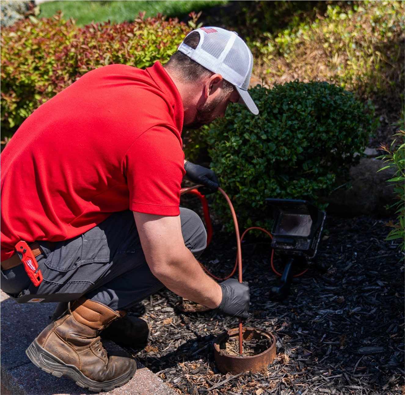 Technician cleaning an outdoor drain near a garden bed.