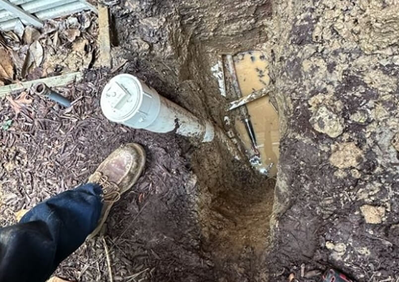 Worker digging in muddy ground to expose underground plumbing.