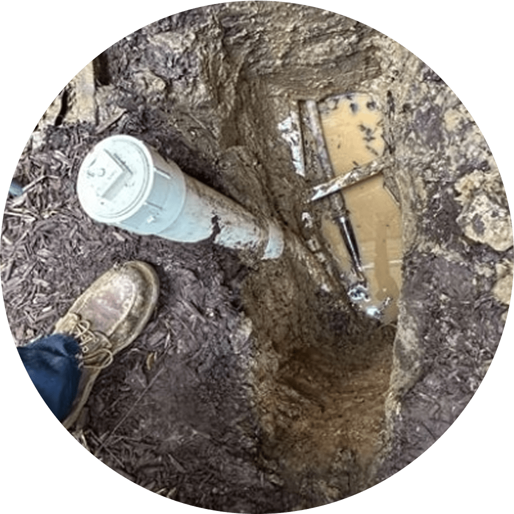Worker digging in muddy ground to expose underground plumbing.