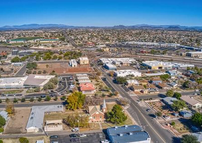 Aerial view of Peoria, Arizona, showing residential neighborhoods, commercial buildings, and tree-lined streets, with distant mountains under a clear blue sky.