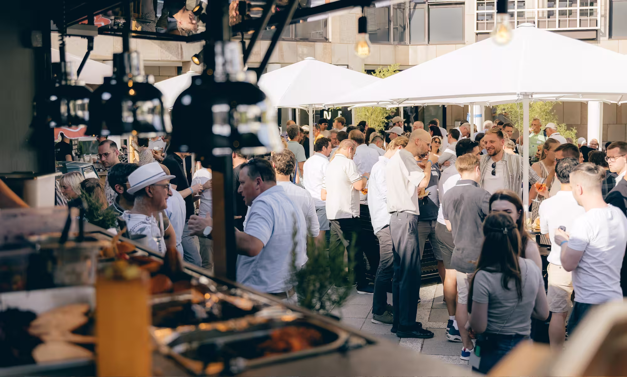 Viele Menschen unter weißen Sonnenschirmen bei einer Firmenfeier im Freien mit Essen und Getränken.