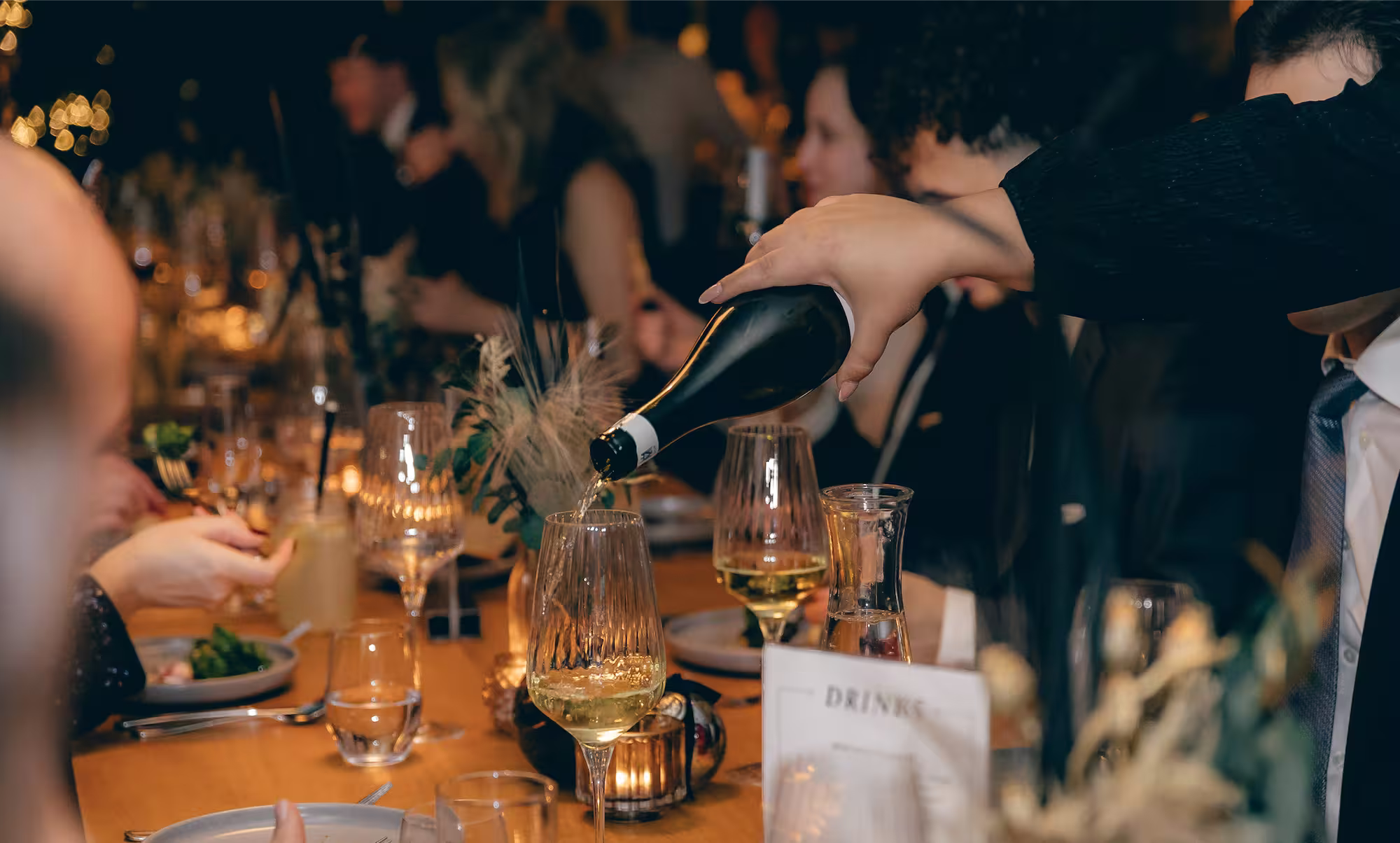 Person gießt weißwein in ein Weinglas bei einem festlichen Abendessen mit mehreren Gästen.