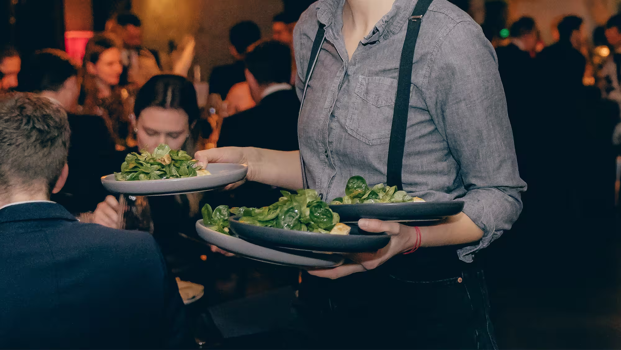 Kellner trägt mehrere Teller mit grünen Salaten in einem belebten Restaurant.