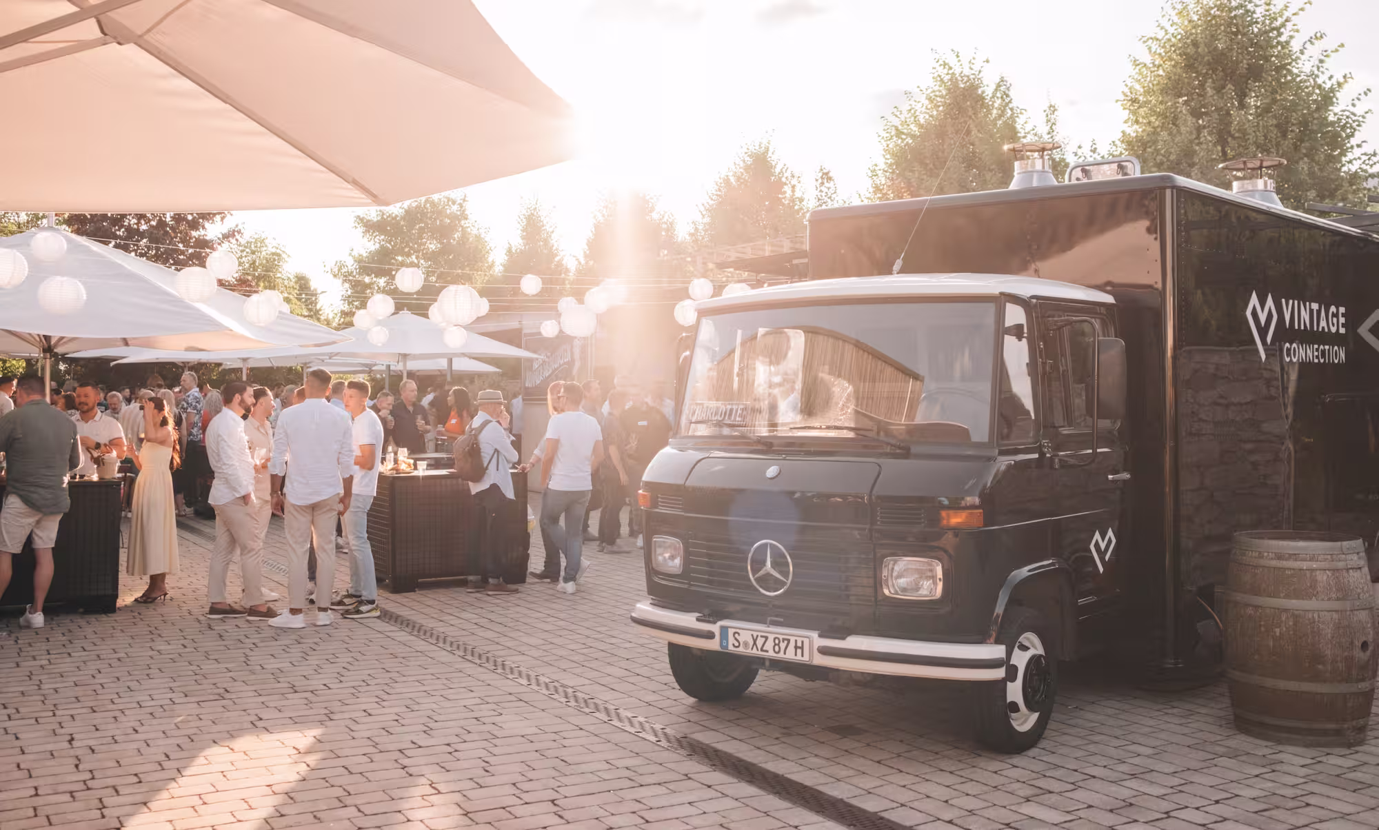 Menschen versammeln sich bei sonnigem Wetter unter Lichterketten und Sonnenschirmen neben einem schwarzen Vintage Mercedes Foodtruck mit Vintage Connection Logo.