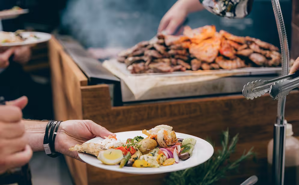 Person hält einen Teller mit mediterranen Speisen vor einem Büfett mit gegrilltem Fleisch und Meeresfrüchten.