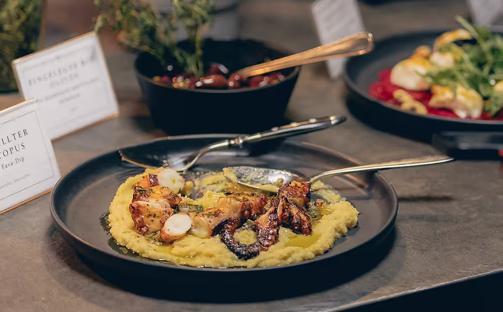 Gegrillter Oktopus serviert auf einer cremigen Fava-Püree mit Löffeln auf einem schwarzen Teller.