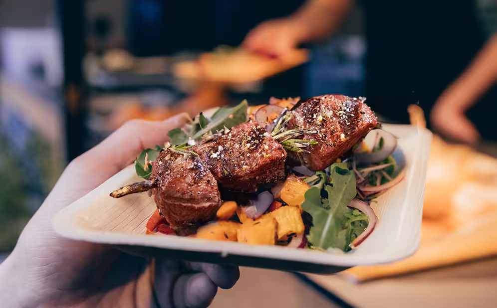Hand hält einen Teller mit einem gegrillten Fleischspieß auf gemischtem Salat mit Rucola, roten Zwiebeln und Würfeln von gegrilltem Gemüse.