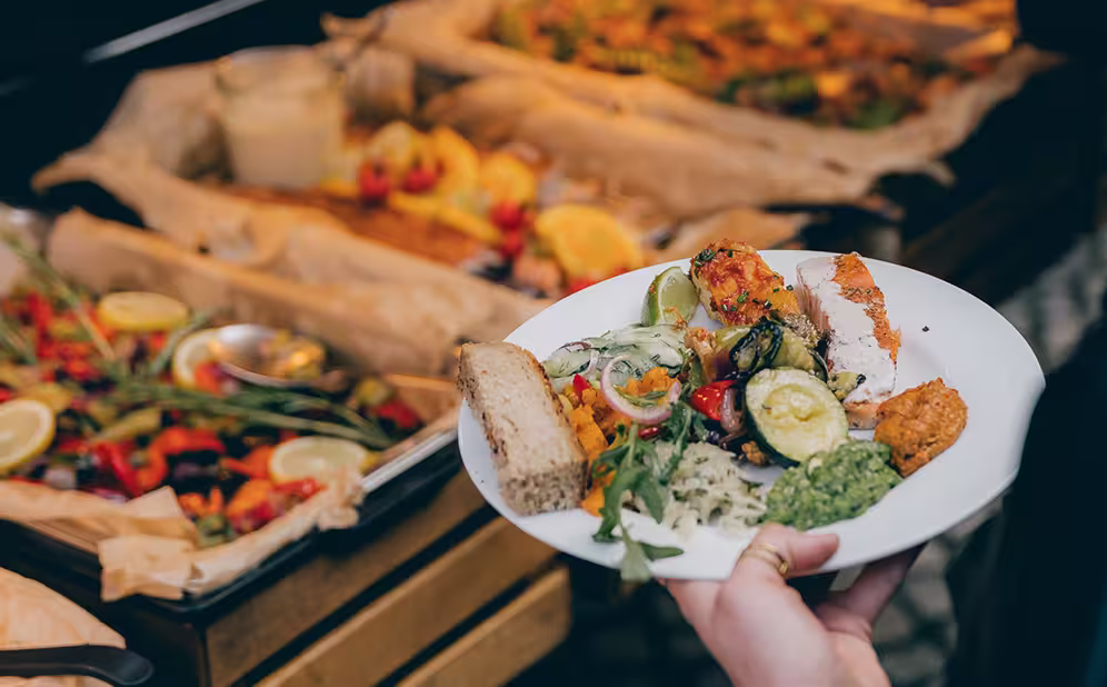 Ein Teller mit verschiedenen Buffetspeisen, darunter Brot, Salat, Zucchini, Lachs und Dips, vor einem Buffet mit belegten Teigböden.