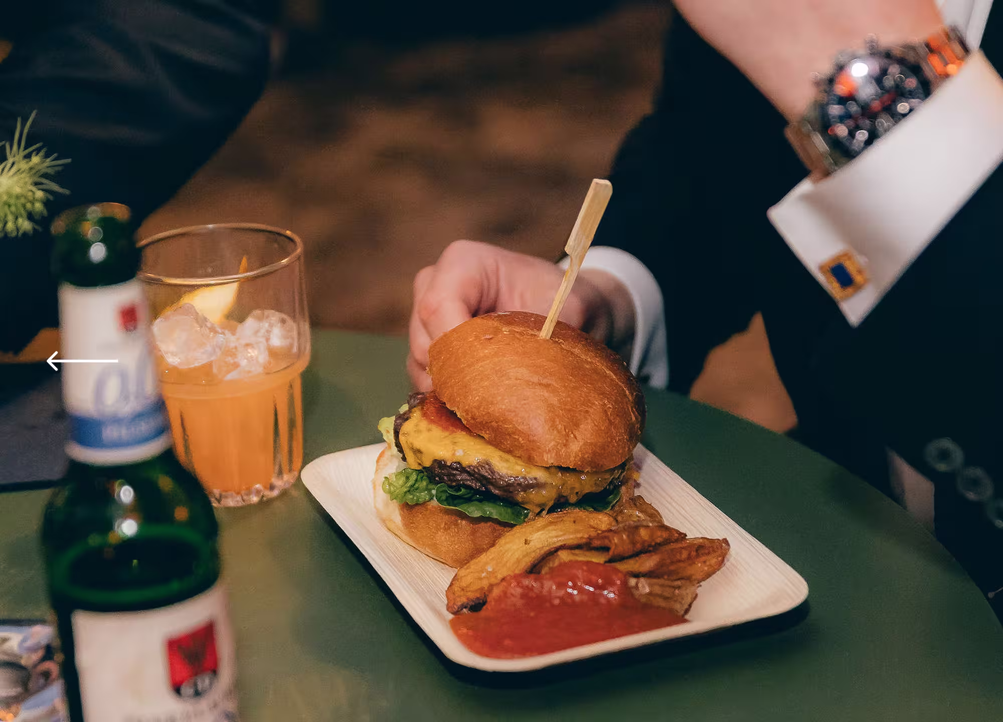 Ein Teller mit einem Cheeseburger, Kartoffelecken und Ketchup neben einem Glas mit Eiswürfeln und einem Getränk auf einem grünen Tisch.