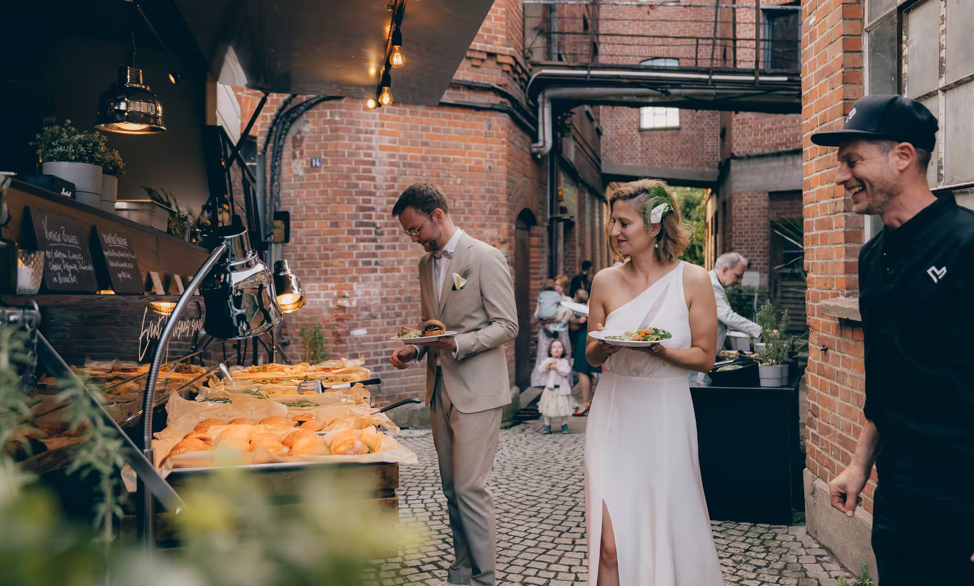 Brautpaar am Buffet mit einem Catering-Mitarbeiter in einem Innenhof mit Backsteinmauern.