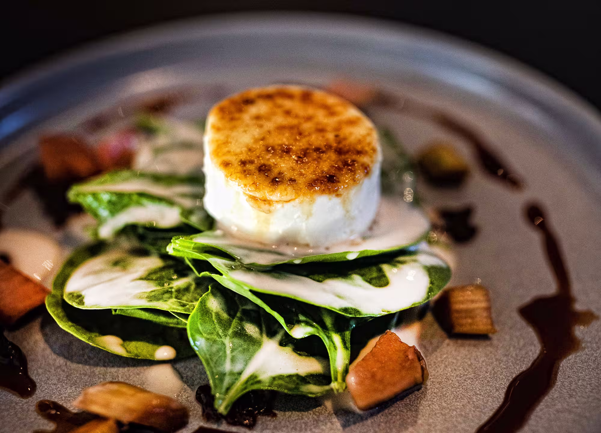 Angerichteter Ziegenkäse mit karamellisierter Oberfläche auf frischem Spinat und cremiger Sauce, garniert mit kleinen Gemüsestücken auf einem grauen Teller.
