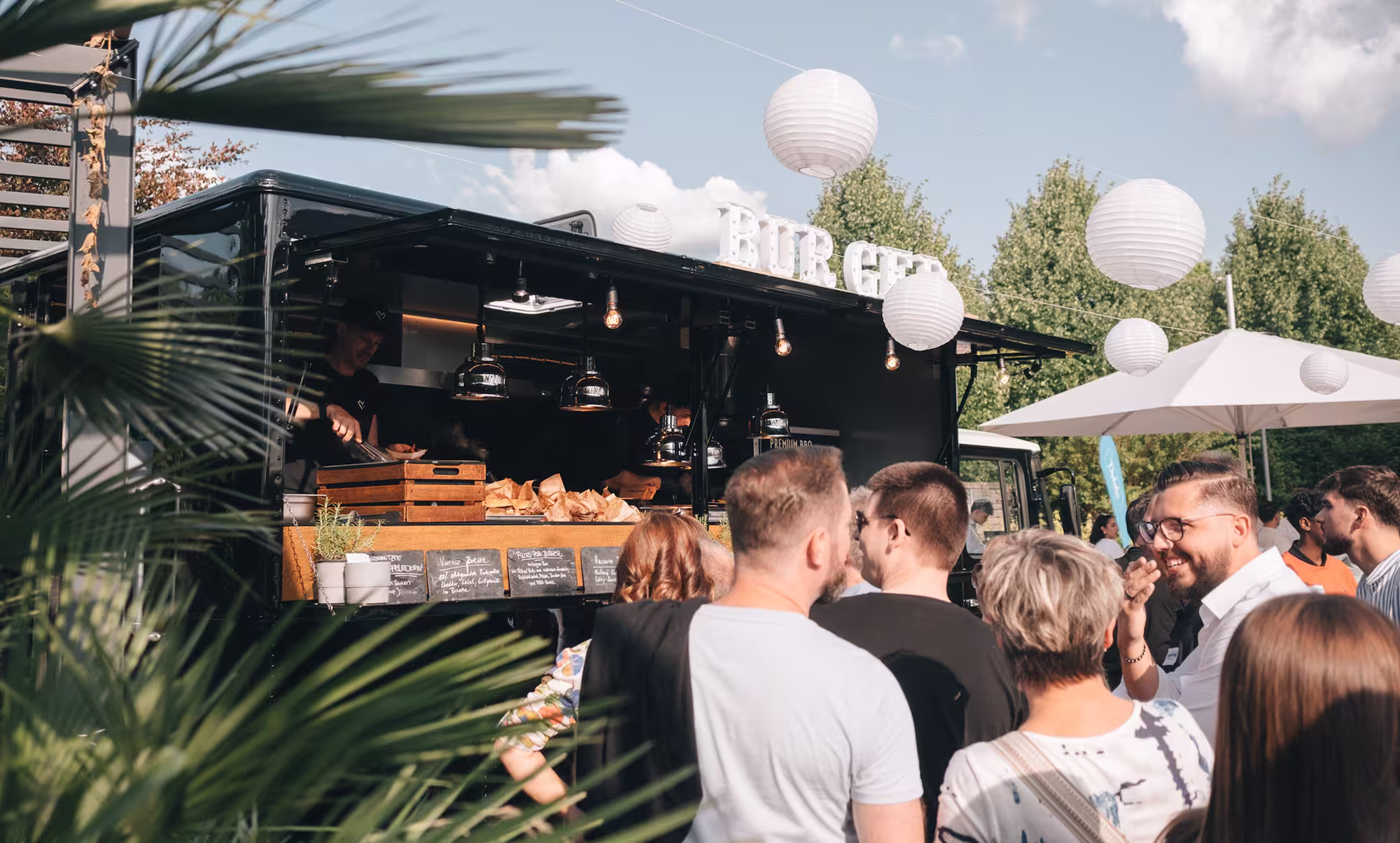 Menschen stehen in einer Schlange vor einem mobilen Burger-Imbissstand mit weißen Laternen und Sonnenschirmen im Freien.