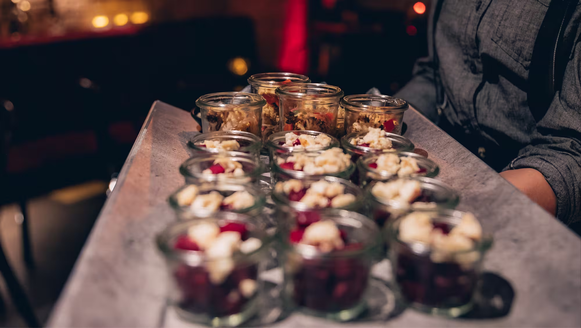 Tablett mit mehreren kleinen Gläsern, gefüllt mit Dessert und Granola, gehalten von einer Person in grauer Kleidung.