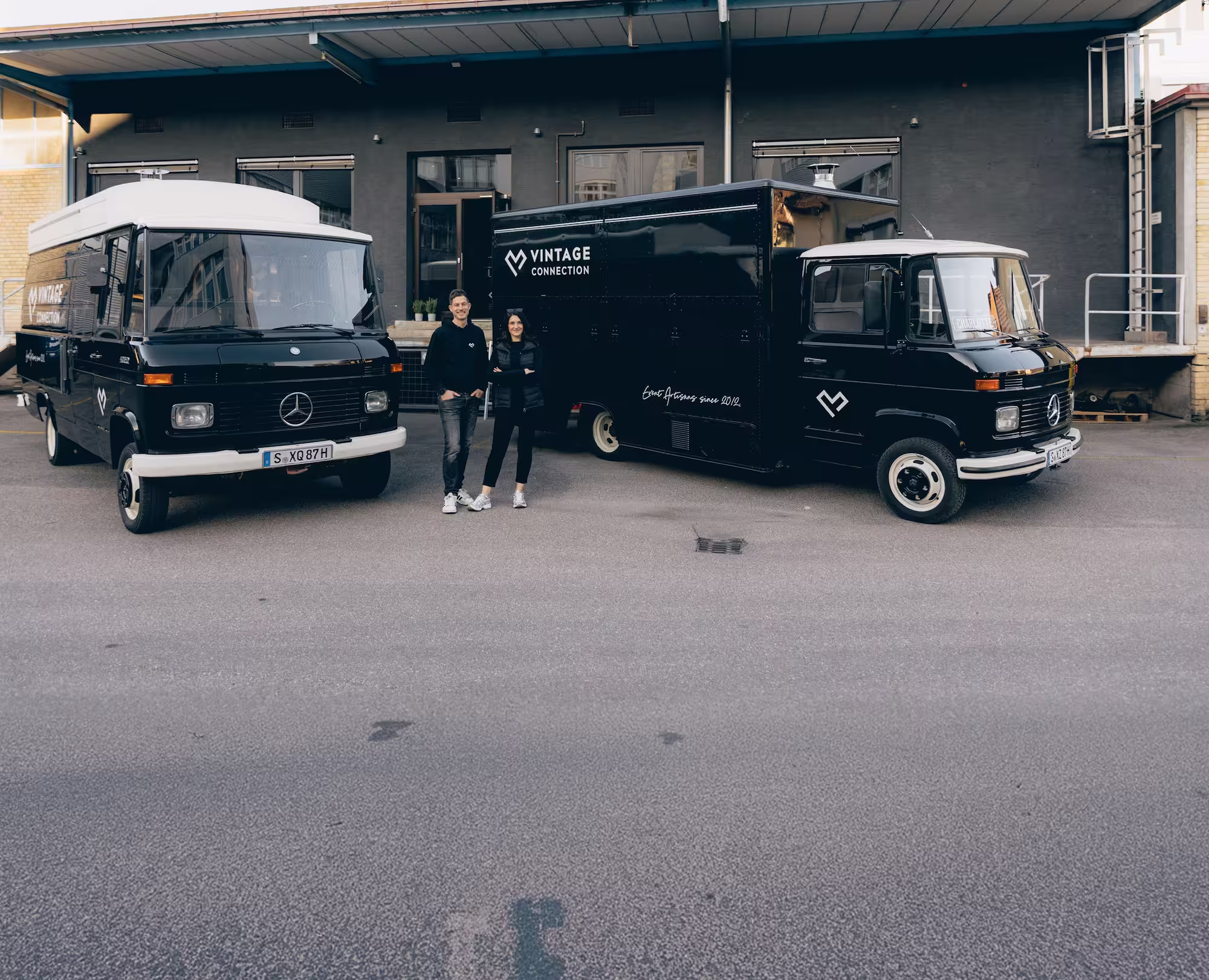 Zwei Personen stehen zwischen zwei schwarzen Vintage Mercedes-Transportern mit dem Schriftzug Vintage Connection vor einem Gebäude.
