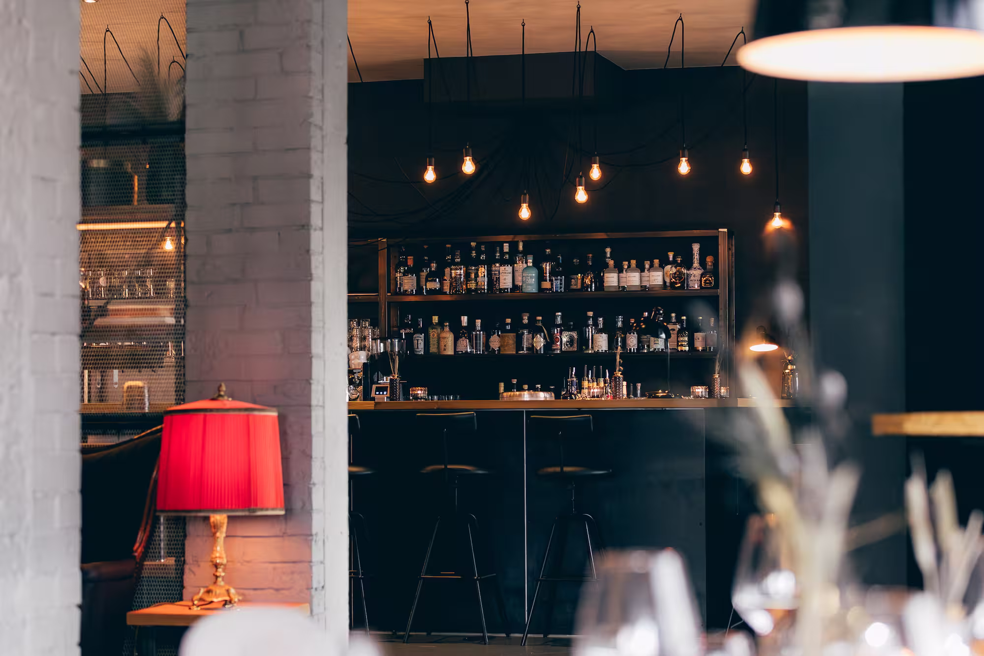 Bar mit schwarzen Hockern, Regalen voller Flaschen und hängenden Glühbirnen in stilvollem Ambiente.