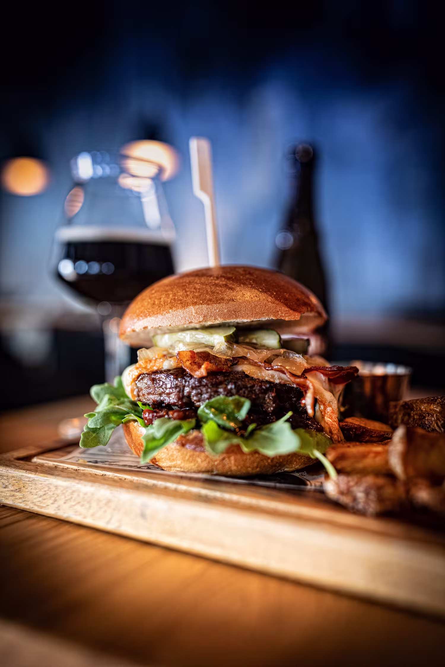 Ein Burger mit Salat, Gurken, Speck und Käse auf einem Holzbrett mit Pommes und dunklem Getränk.