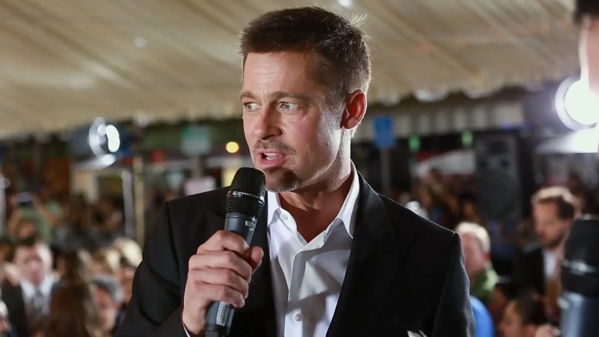 Man in a black suit and white shirt speaking into a microphone at a crowded indoor event.