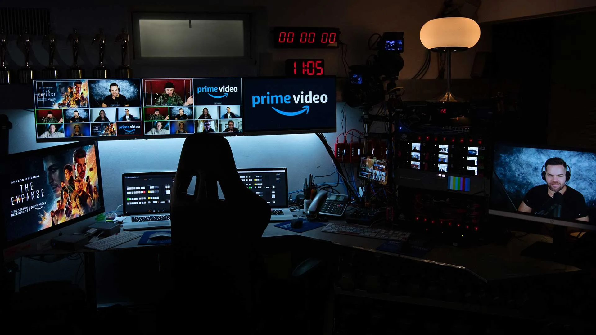Dimly lit broadcast control room with multiple monitors showing Prime Video content and a video call grid, a digital clock reading 11:05, and a person on a headset visible on one screen.