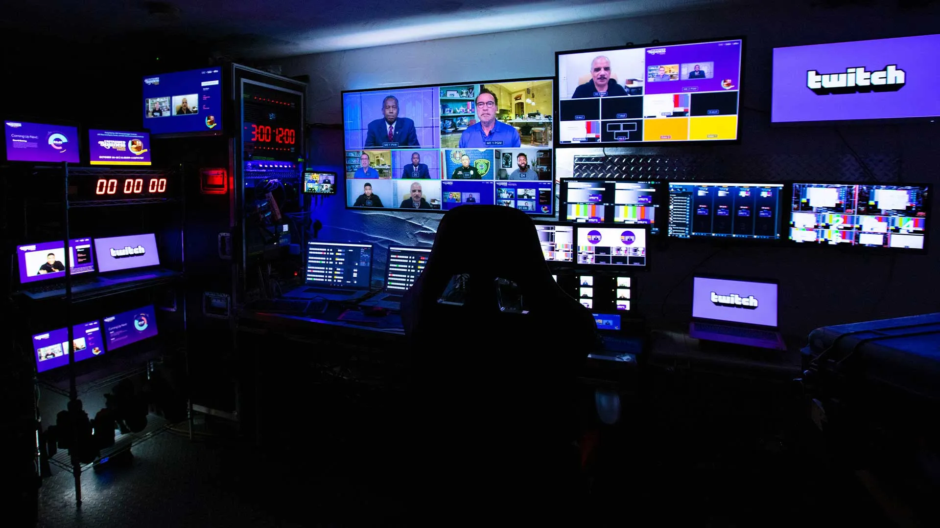 Dark control room with multiple monitors displaying video call participants and Twitch branding.