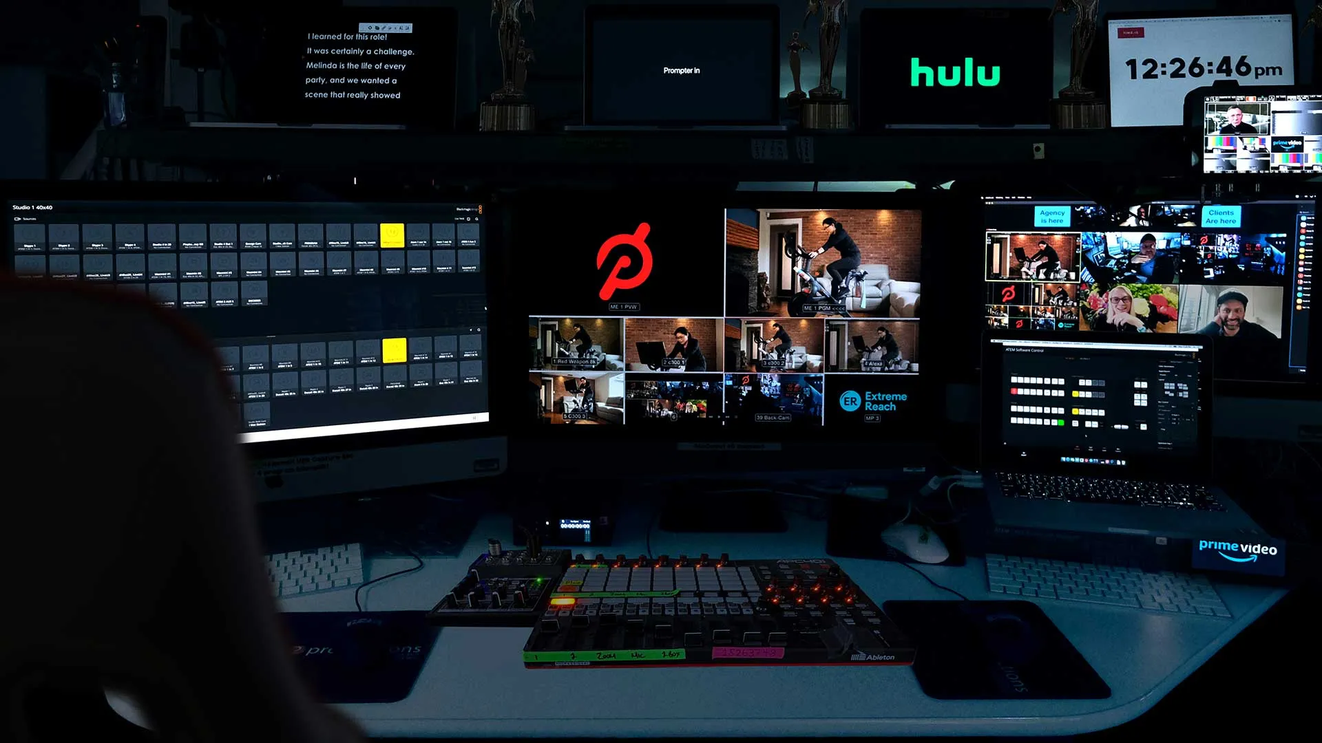 A dimly lit video production control room with multiple screens showing live camera feeds, a Hulu logo, Prime Video interface, and video editing controls on a desk.