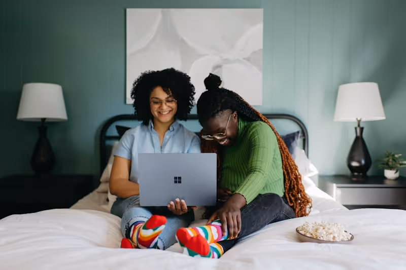 Two women sitting on a bed looking at a laptop and smiling, both wearing colorful striped socks.