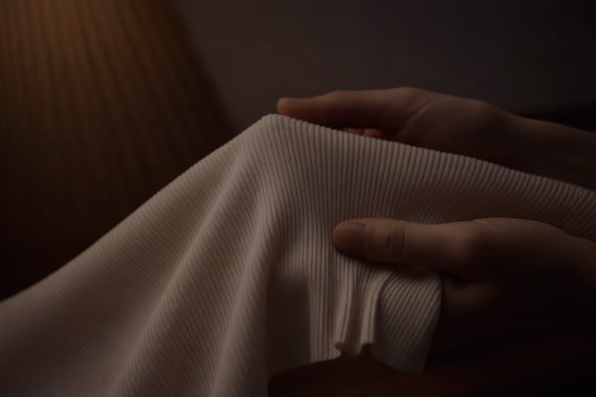 Two hands gently holding a piece of light-colored ribbed fabric against a dark background.