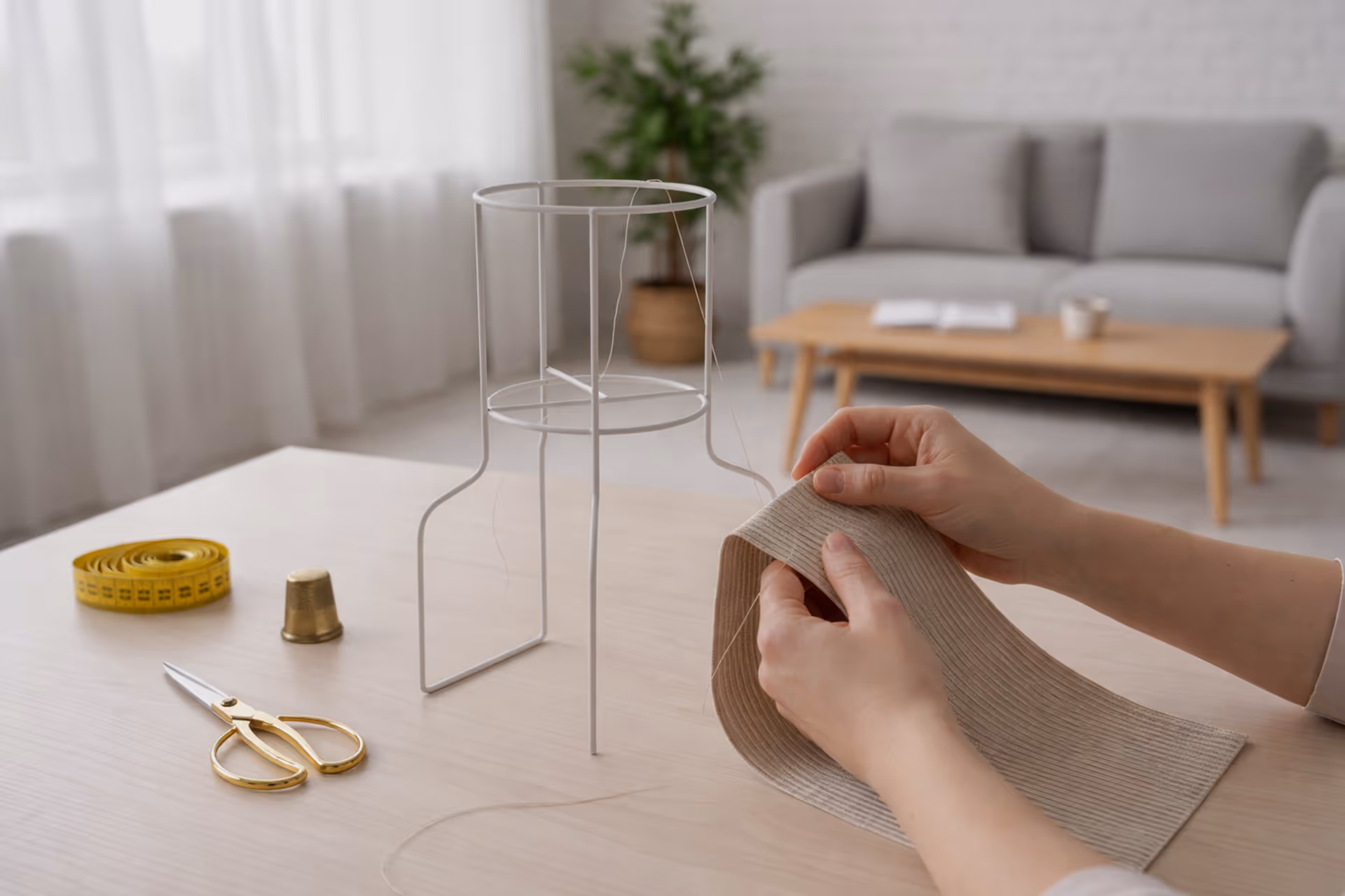 Hands sewing beige ribbed fabric next to a metal lampshade frame, golden scissors, yellow measuring tape, and thimble on a wooden table.
