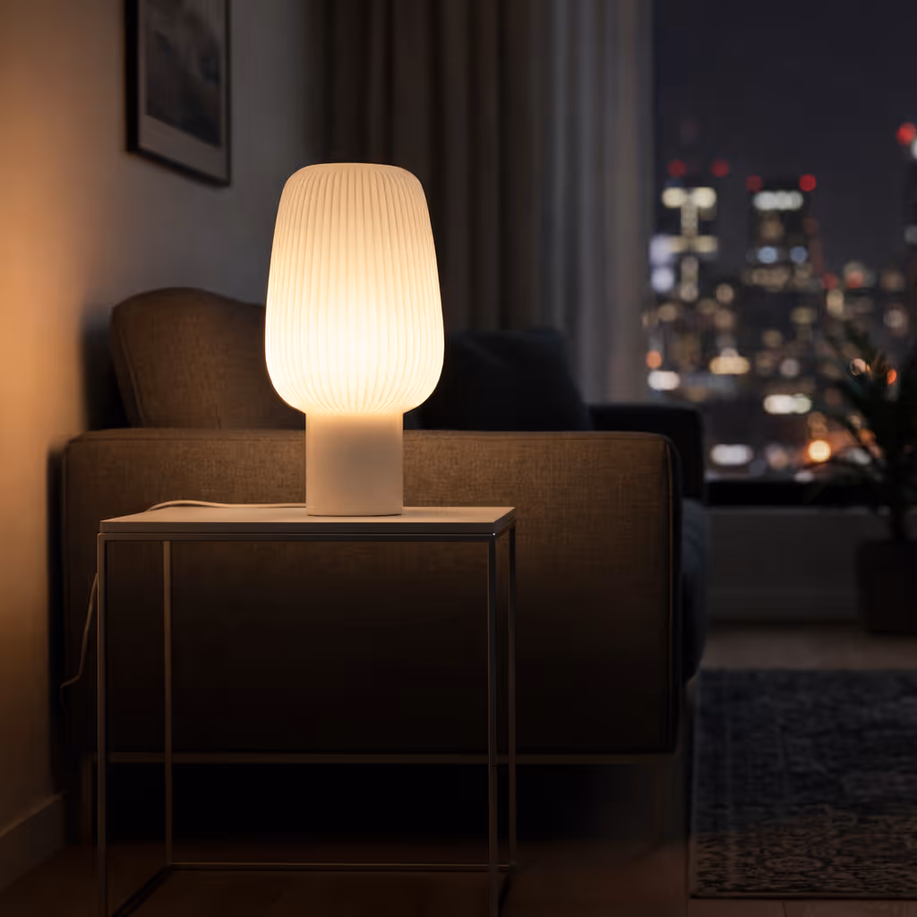 Modern table lamp with a ribbed white shade glowing warmly on a side table next to a brown sofa in a dimly lit living room at night.