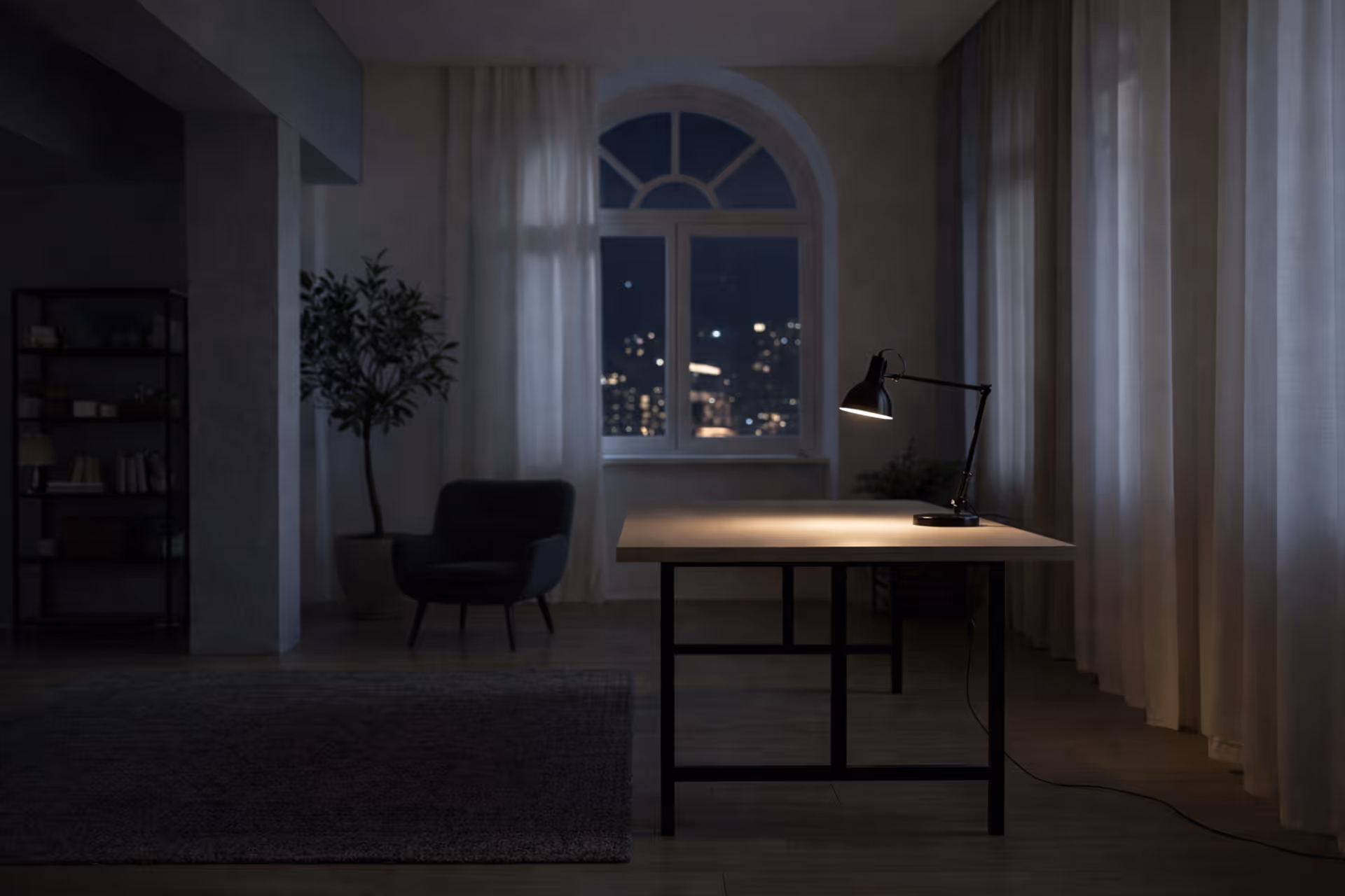 Dimly lit modern room with a wooden table illuminated by a desk lamp, a dark armchair, a tall plant, and a window showing city lights at night.