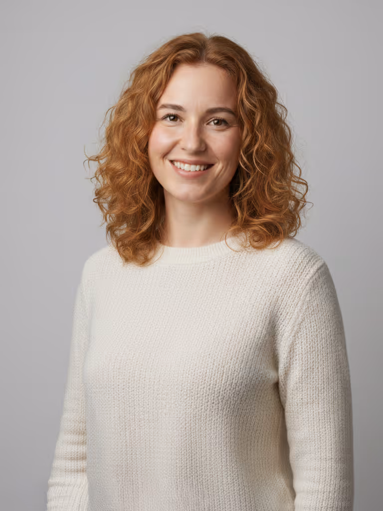 Smiling young woman with curly red hair wearing a white knitted sweater against a gray background.