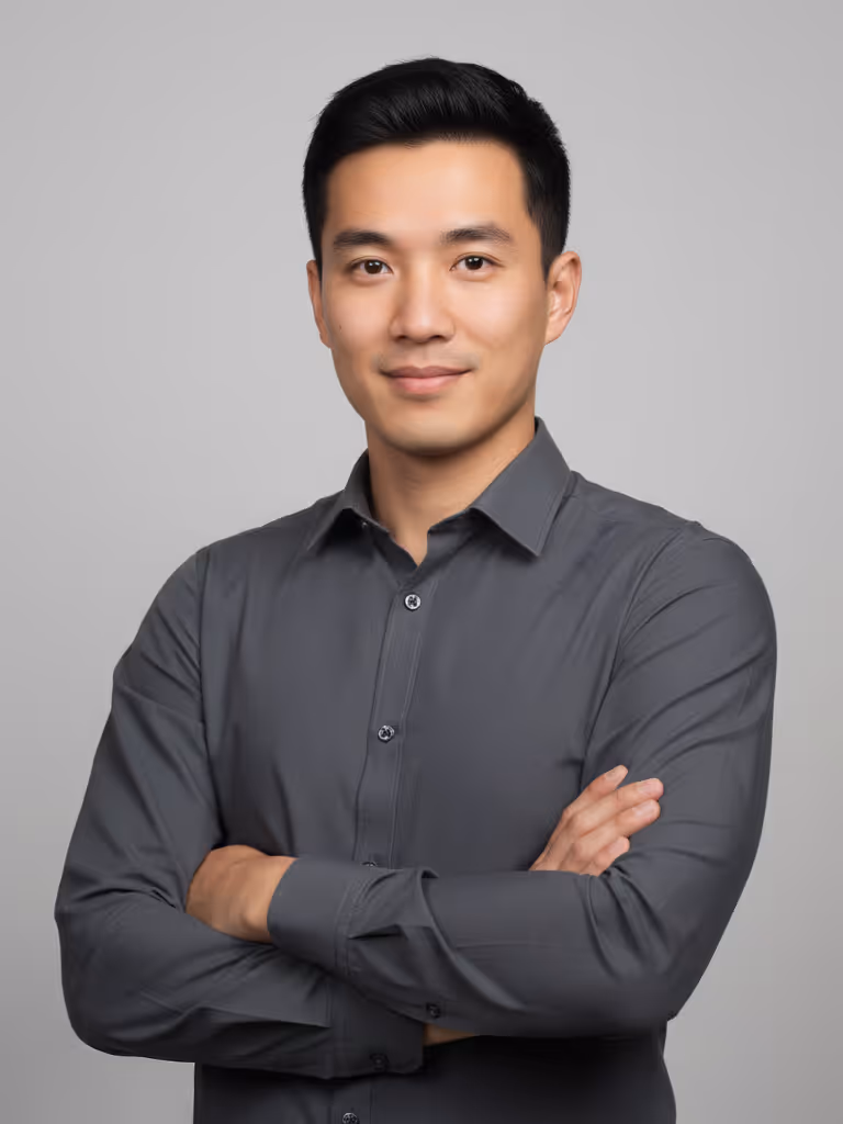 Young man with short black hair wearing a dark gray shirt, standing with arms crossed and smiling slightly against a plain gray background.