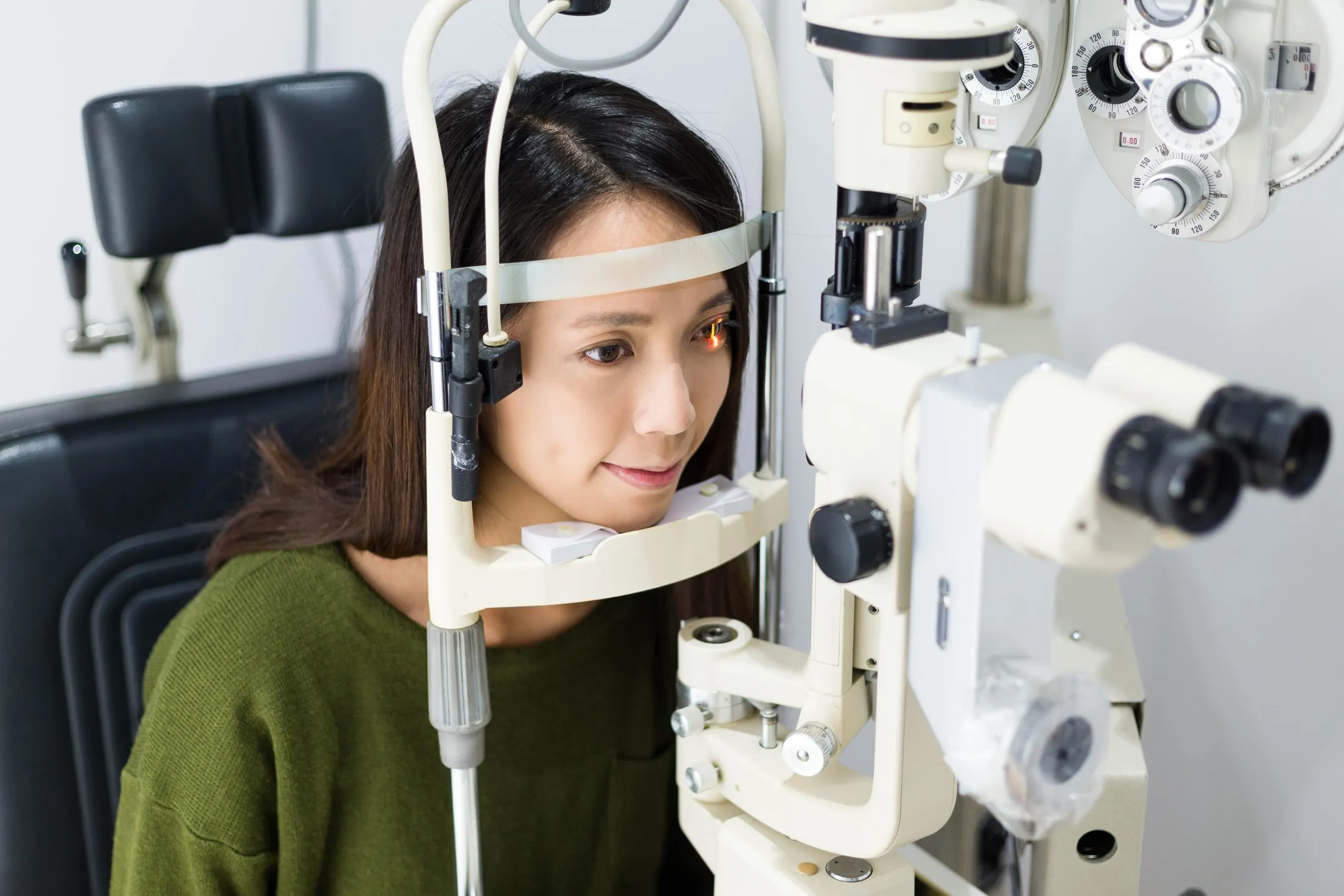 Woman wearing green sweater undergoing eye exam using slit lamp exam machine.