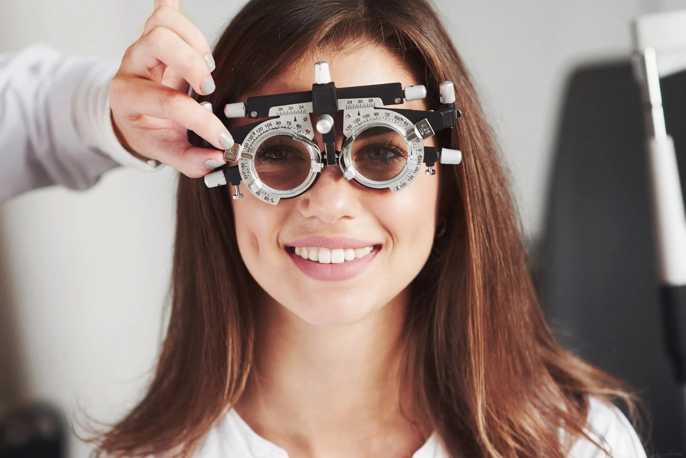 Smiling woman wearing an eye exam phoropter held by an optometrist’s hand.