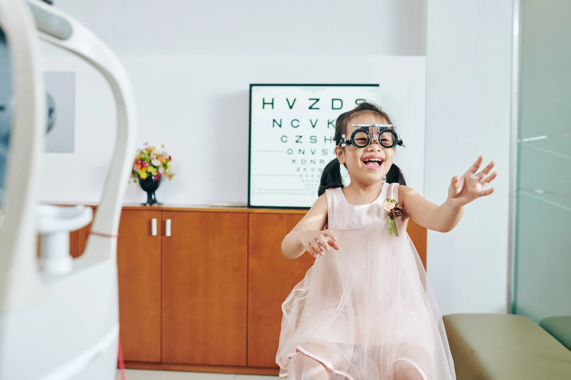 Smiling young girl wearing eye test glasses in an optometrist office with eye chart behind her.